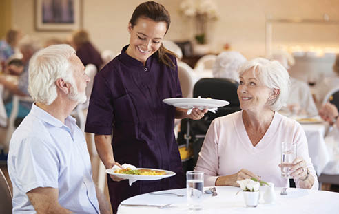 Senior Couple Being Served With Meal By Carer In Dining Room Of Retirement Home