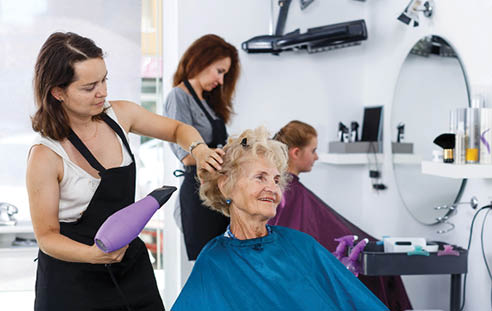Professional female stylist making hairdo for senior woman in salon, using hair dryer 